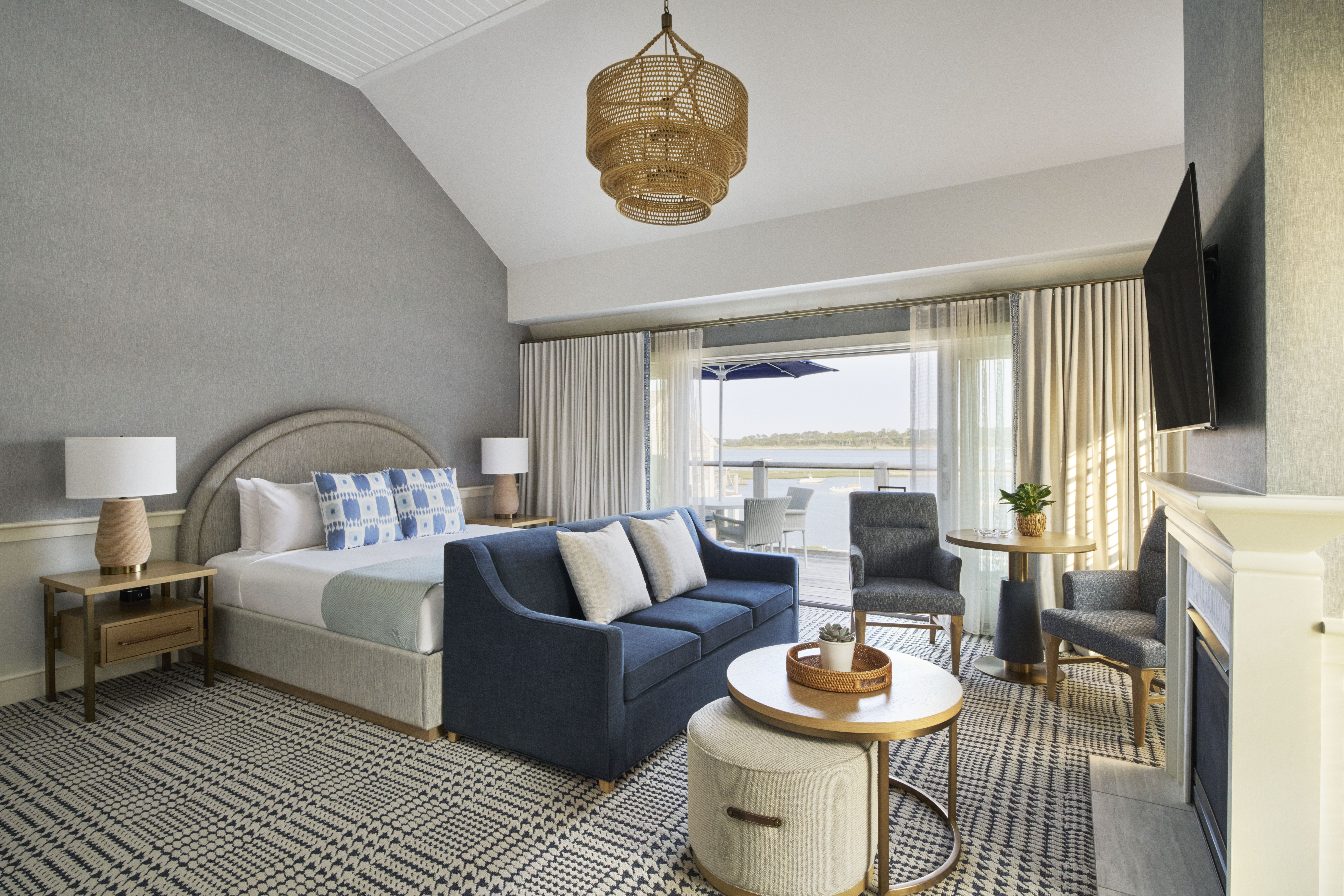 A modern hotel room at Wequassett Resort and Golf Club features a king bed, blue sofa, round coffee table, small dining set, and large windows overlooking a balcony with a stunning water view at this premier Cape Cod resort.