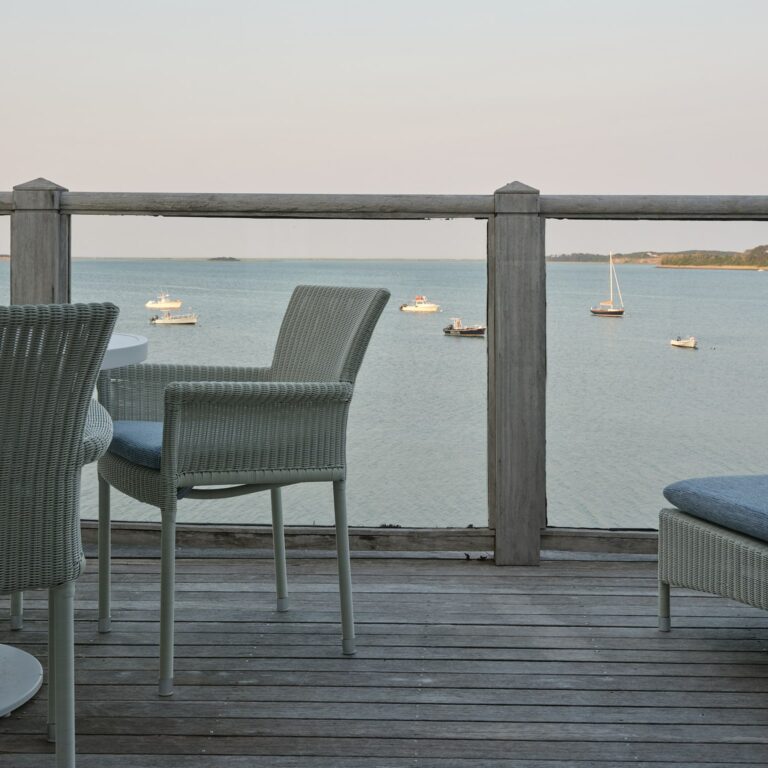 A wooden deck with wicker chairs overlooks a calm body of water with several anchored boats visible in the distance, offering a serene setting ideal for a Cape Cod wedding venue.