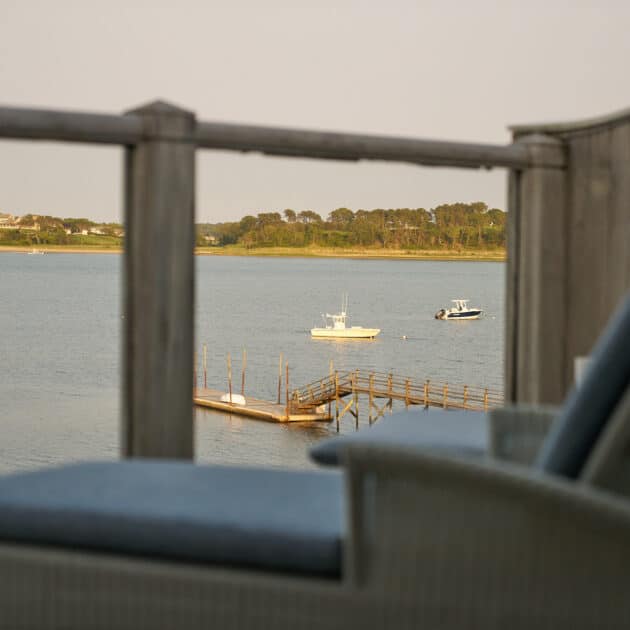 View of a calm body of water with two boats, a wooden dock, and distant trees, seen from behind a balcony railing and outdoor lounge chairs.