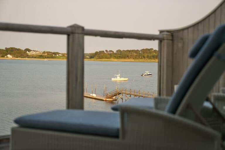 View of a calm body of water with two boats, a wooden dock, and distant trees, seen from behind a balcony railing and outdoor lounge chairs.