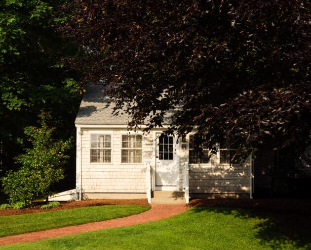 Charming small white house with a shingle exterior, white-framed windows, and a front porch—just minutes from 28 Atlantic and ideal as a Cape Cod wedding venue. Curved brick path leads to the door, shaded by large trees.