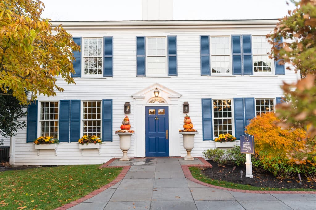 White two-story house with blue shutters, blue front door, fall flowers, and pumpkins stacked in urns by the entrance—creating a warm, inviting atmosphere reminiscent of Fall Festivals. Plaque sign is visible on the right side near a small tree.