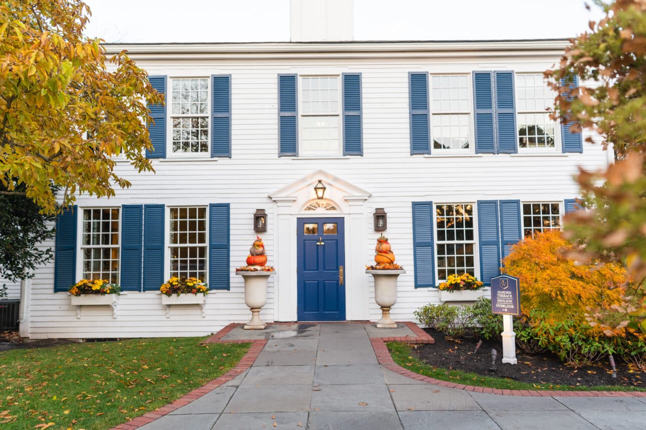 White two-story house with blue shutters, blue front door, fall flowers, and pumpkins stacked in urns by the entrance—creating a warm, inviting atmosphere reminiscent of Fall Festivals. Plaque sign is visible on the right side near a small tree.