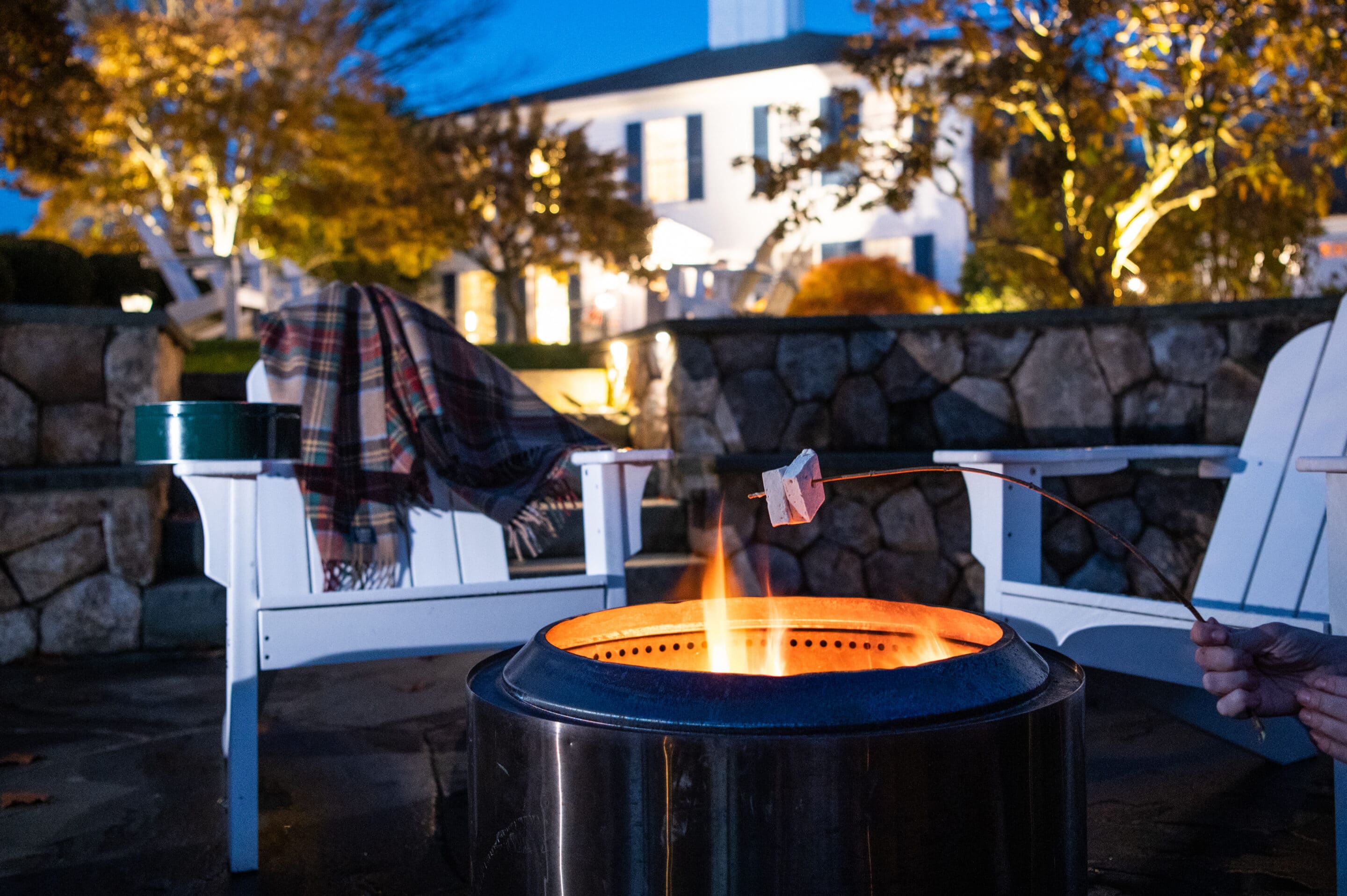 A hand roasts a marshmallow over a fire pit near two white chairs; a plaid blanket rests on one chair, with a house and trees visible in the background at dusk.