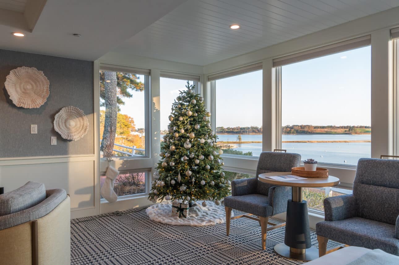 A Christmas tree with silver ornaments stands in the corner of a room beside large windows overlooking a bay, where seasonal deals and special offers await.