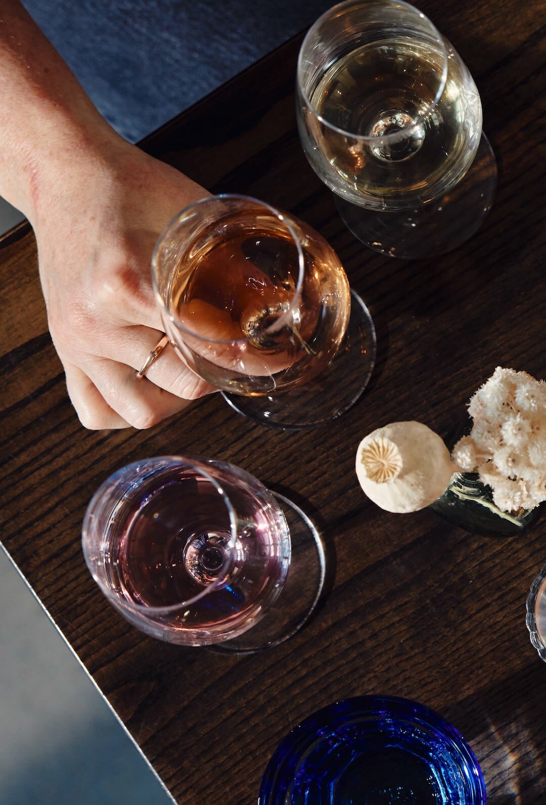 Overhead view of two people holding wine glasses at a wooden table during a Wine Dinner, with several other glasses, blue water cups, a lit candle, and a small floral decoration.