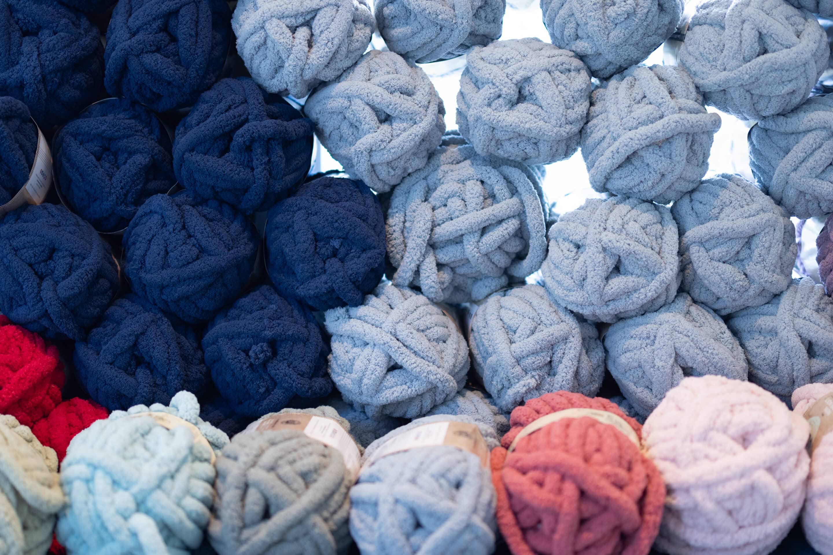 Assorted skeins of chunky yarn in blue, red, pink, and light blue colors are stacked closely together in rows, perfect for creating your next Blanket Pattern or mastering the Herringbone Stitch at your upcoming Blanket Party.