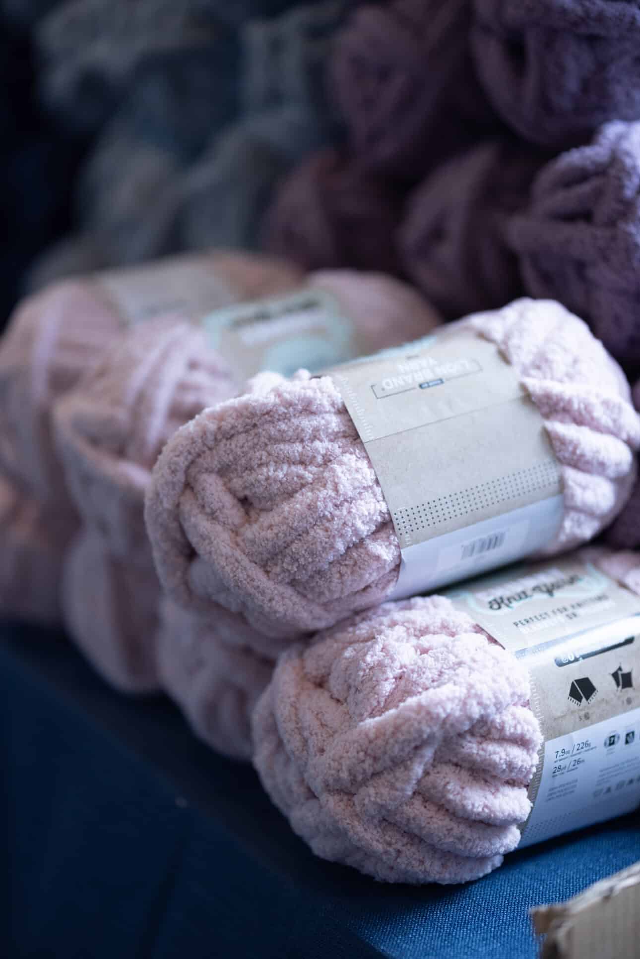 Three skeins of bulky, pale pink yarn with labels are stacked on a blue surface—perfect for your next herringbone stitch project. In the background, other yarn in various colors adds inspiration for a true blanket party.