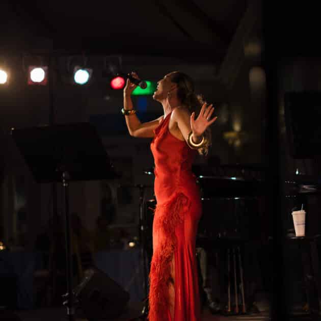 A woman in a red dress sings passionately on stage at the Cape Cod Jazz Festival, holding a microphone, with stage lights behind her and a music stand nearby.