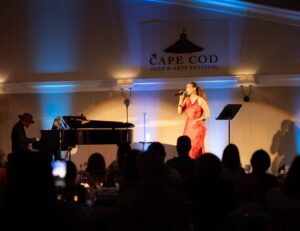 A singer performs on stage with a pianist at the Cape Cod Jazz & Arts Festival during Wequassett evenings, while an audience watches in a dimly lit venue.