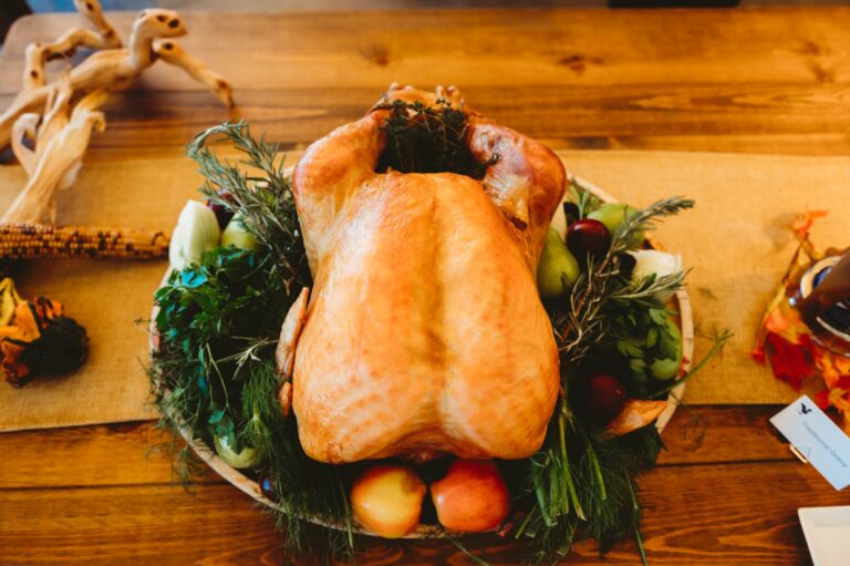 A roasted whole turkey on a platter garnished with herbs, fruits, and vegetables, placed on a wooden table.