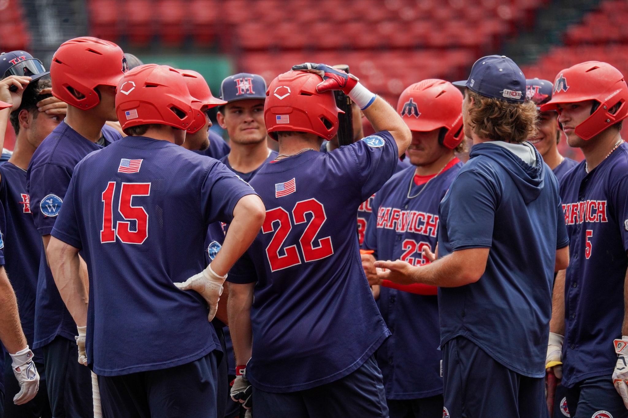 A group of Harwich Mariners baseball players wearing red helmets and blue uniforms with numbers gather in a huddle on the field.