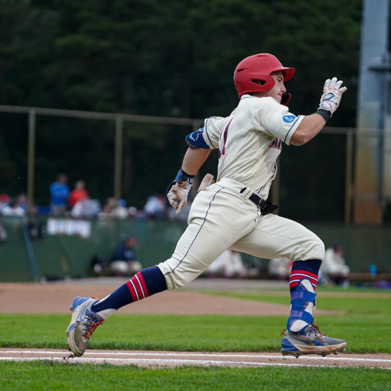 A baseball player in a white uniform and red helmet runs toward first base during a game, with the field and other players in the background.