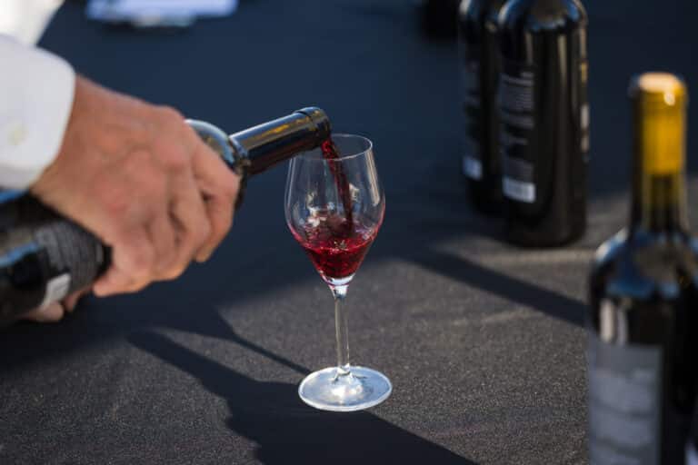 A person pours red wine from a Harvest Reds bottle into a wine glass on a table, with other wine bottles in the background.