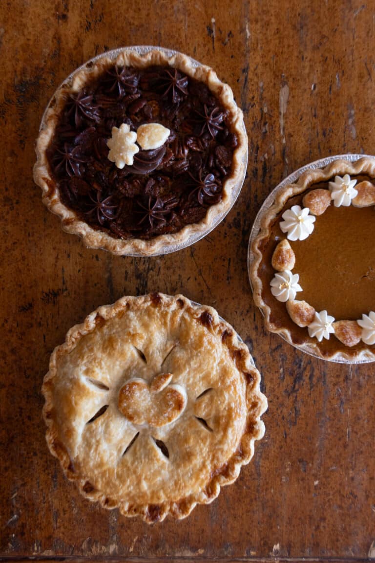 Three different pies sit on a wooden table: a chocolate pie with dark swirls, a pumpkin pie with floral decorations, and a fruit pie with a heart-shaped crust design.