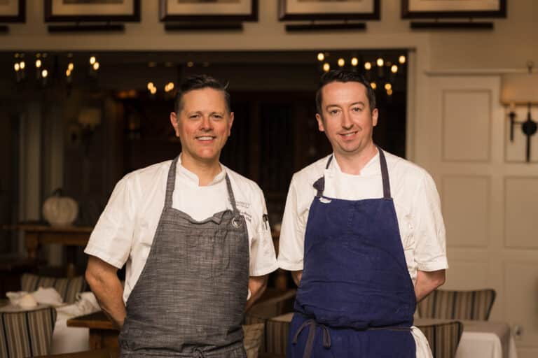 Two male chefs stand side by side in the Wequassett restaurant dining area, both wearing white shirts and aprons, smiling at the camera—ready to welcome guests to their Supper Club experience.