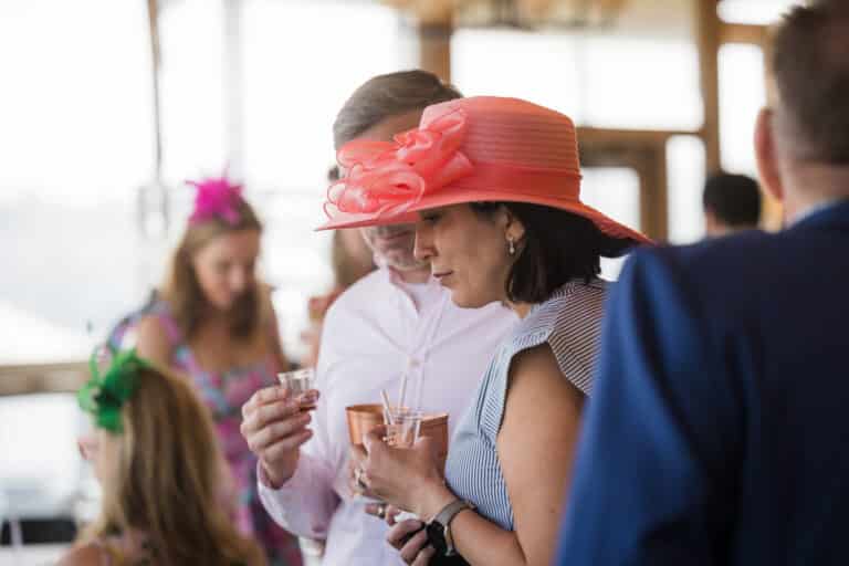 A woman in a large pink hat and a man in a white shirt hold drinks and chat at a Kentucky Derby-inspired social event, with other guests mingling in the background.