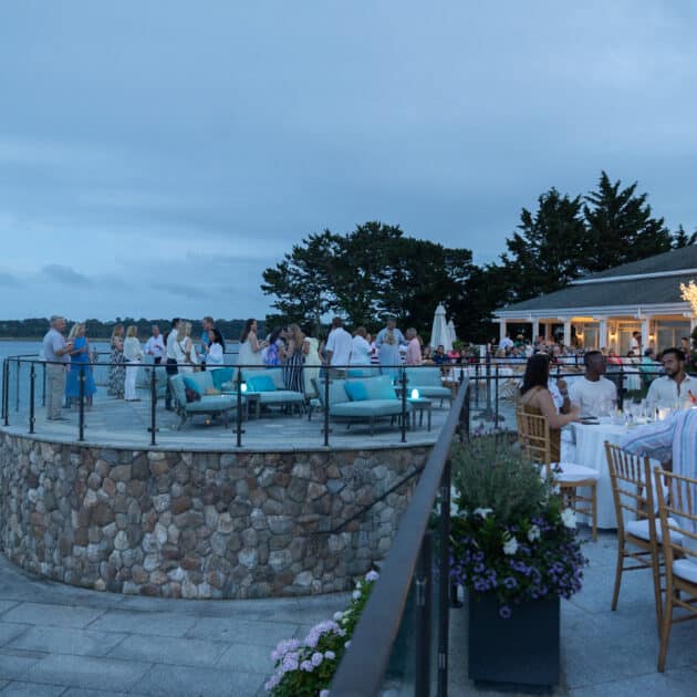 People sit at tables and gather on a stone terrace overlooking the water at an outdoor evening Jazz Festival near a building on Cape Cod, beautifully decorated with lights and flowers.