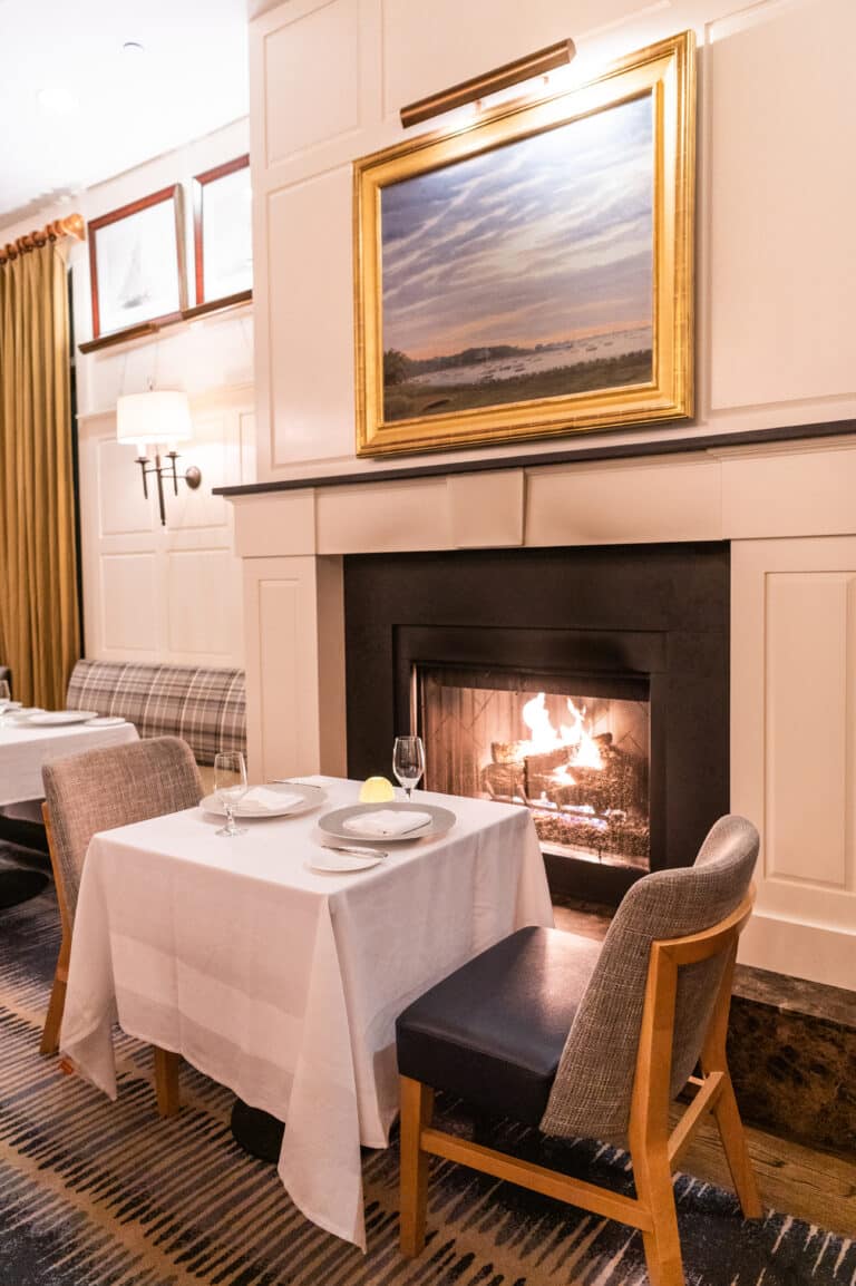 A small dining table for two with white tablecloth is set in front of a lit fireplace, beneath a framed landscape painting in an elegant restaurant interior.
