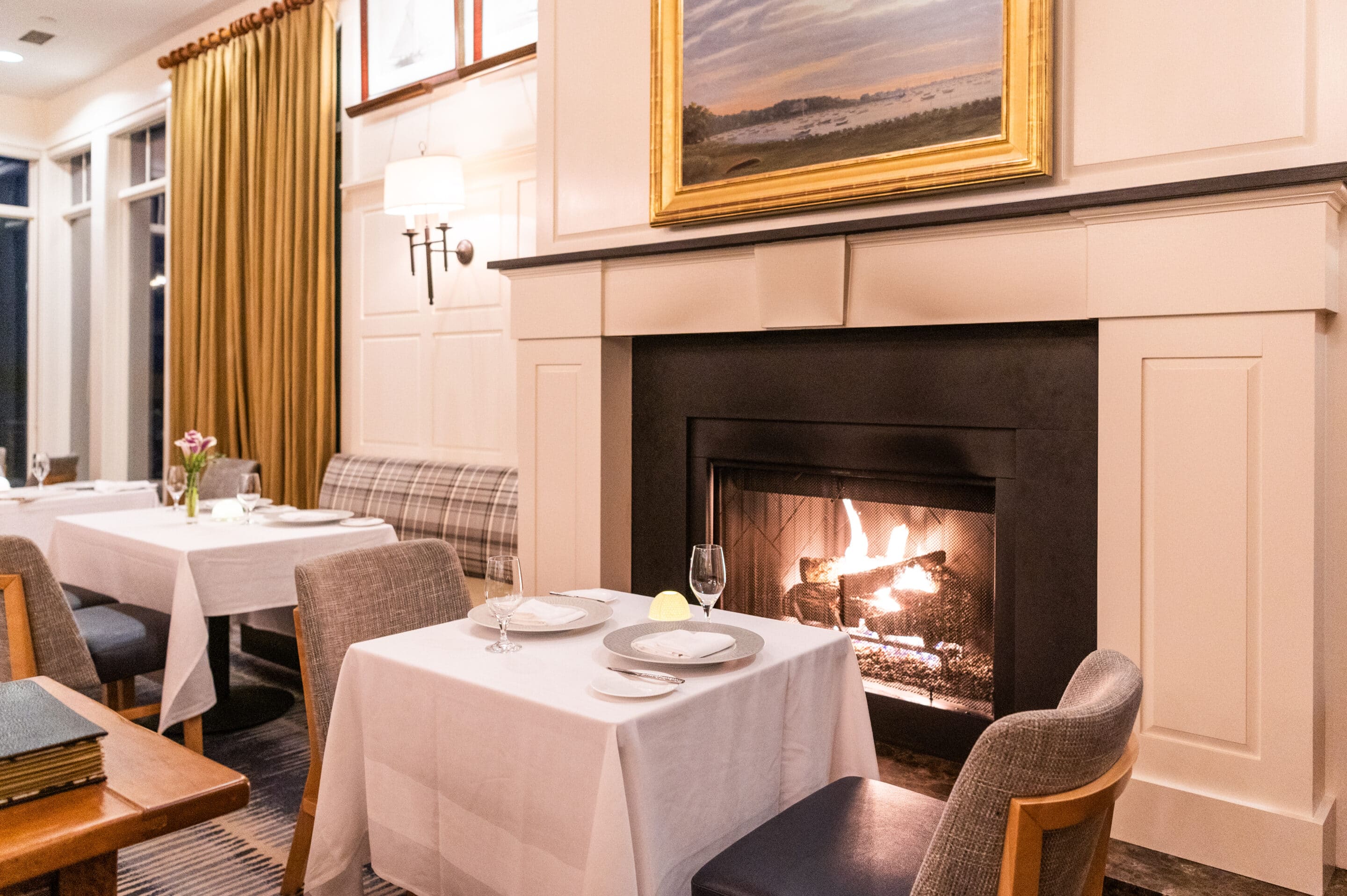 A cozy restaurant dining area with white tablecloths, set tables, upholstered chairs, and a lit fireplace beneath a landscape painting sets the scene for an intimate evening after enjoying Cape Symphony Presents at Wequassett.