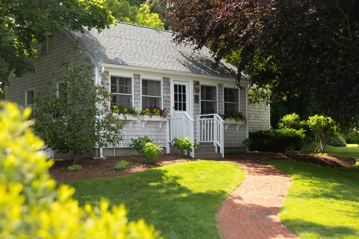 Small grey Tulip Cottage with white trim, flower boxes under the windows, and a brick walkway leading to the front steps, surrounded by green lawn and trees on a sunny day.