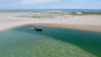 A small boat embarks on a journey through clear green water near a sandy beach with dunes under a partly cloudy sky.