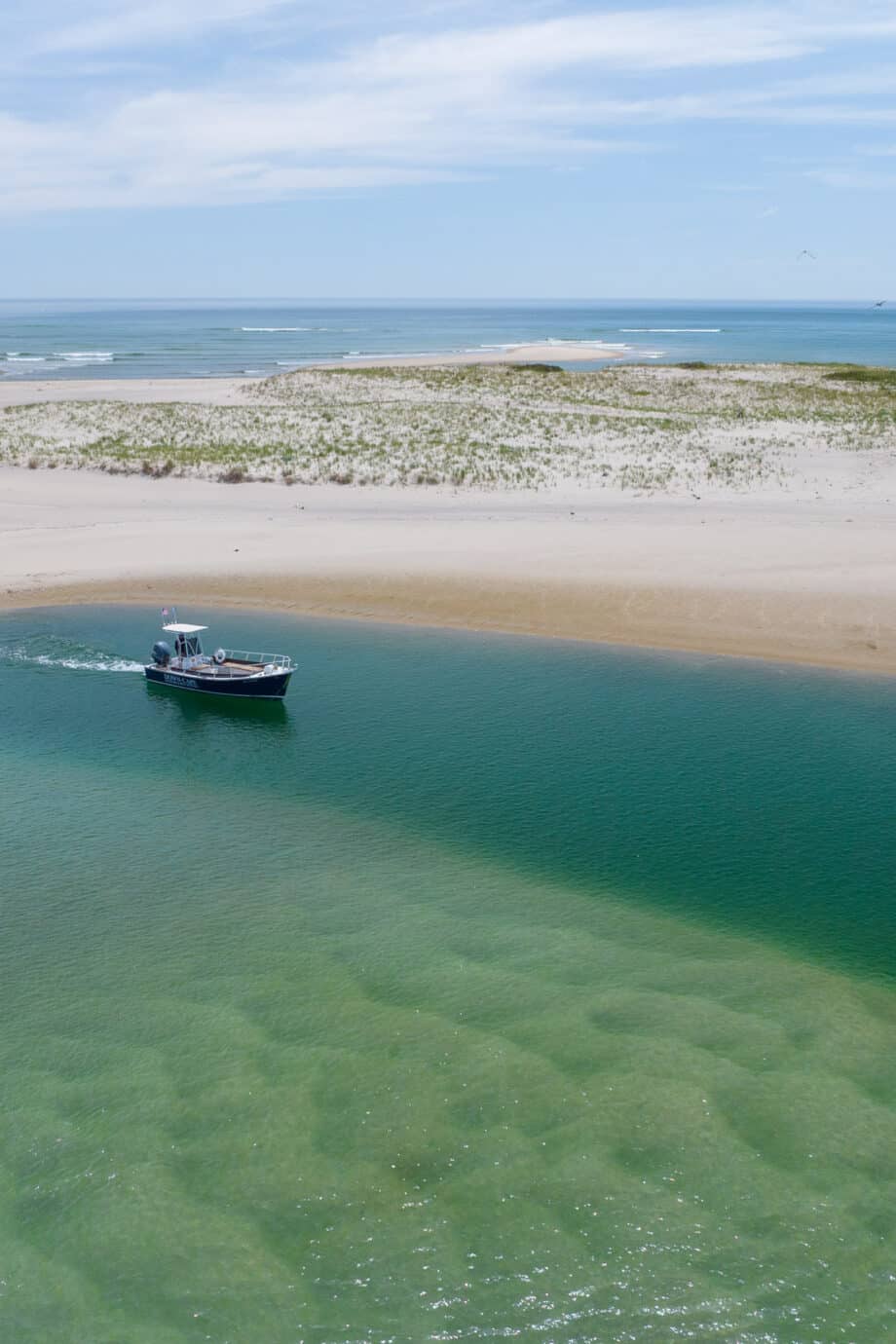 A small boat embarks on a journey through clear green water near a sandy beach with dunes under a partly cloudy sky.