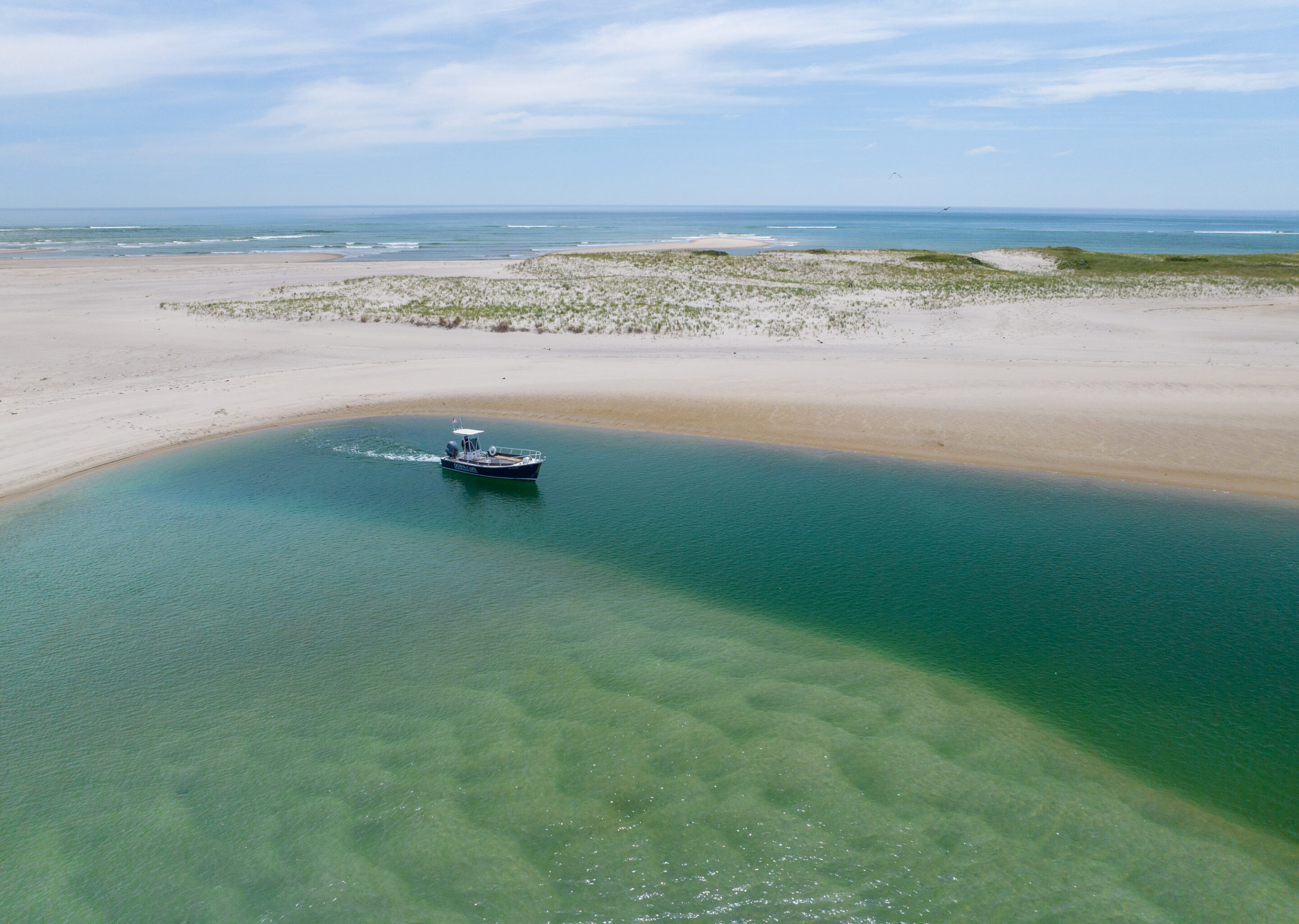 A small boat embarks on a journey through clear green water near a sandy beach with dunes under a partly cloudy sky.