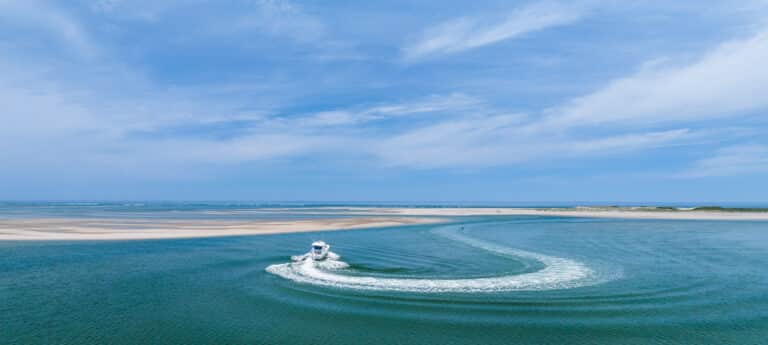 A small motorboat makes a wide curved wake on calm blue water near a sandy coastline under a partly cloudy sky, offering unforgettable On the Water Experiences.