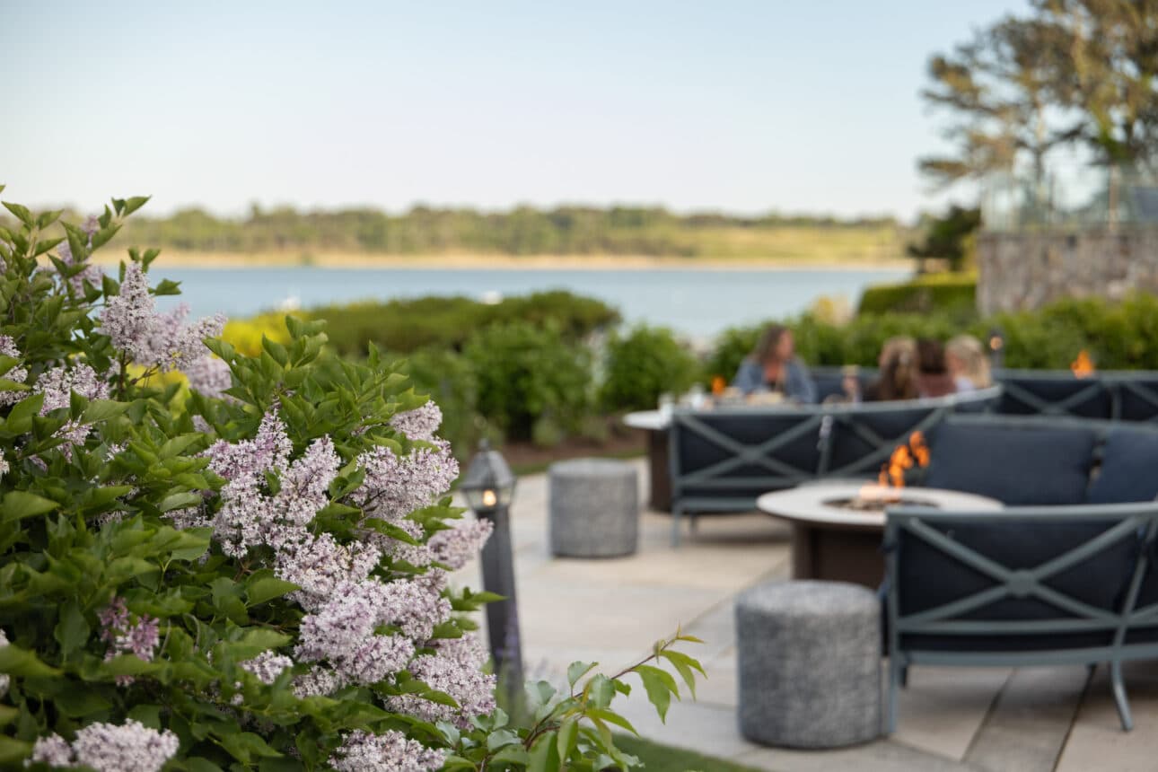 The Verandahs' outdoor patio features lilac bushes in the foreground, blue cushioned seating, fire pits, and people talking near a waterfront on a clear day.