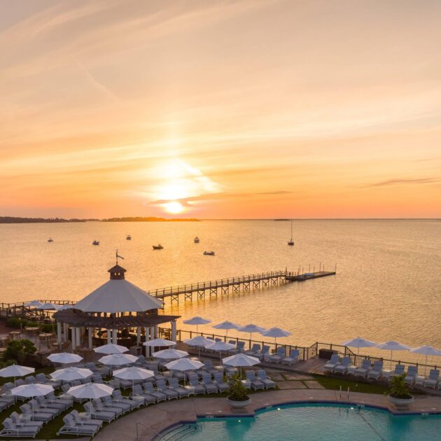 Resort pool area with lounge chairs and umbrellas overlooking a calm bay at sunset; evenings at Wequassett are serene, with a pier extending into the water and boats anchored nearby.