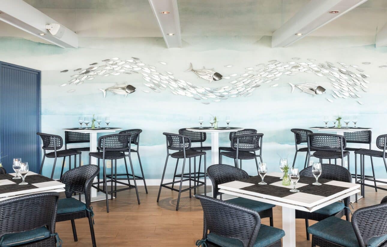 Modern restaurant with wicker chairs, tall tables, and a wall mural featuring metal fish art on a blue gradient background. Wine glasses and place settings are arranged on the tables.
