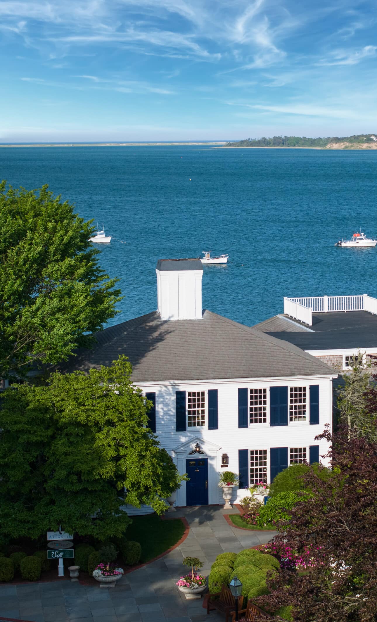 White house with blue shutters and a cupola overlooks a calm blue bay, with several small boats on the water and a distant shoreline under a partly cloudy sky.