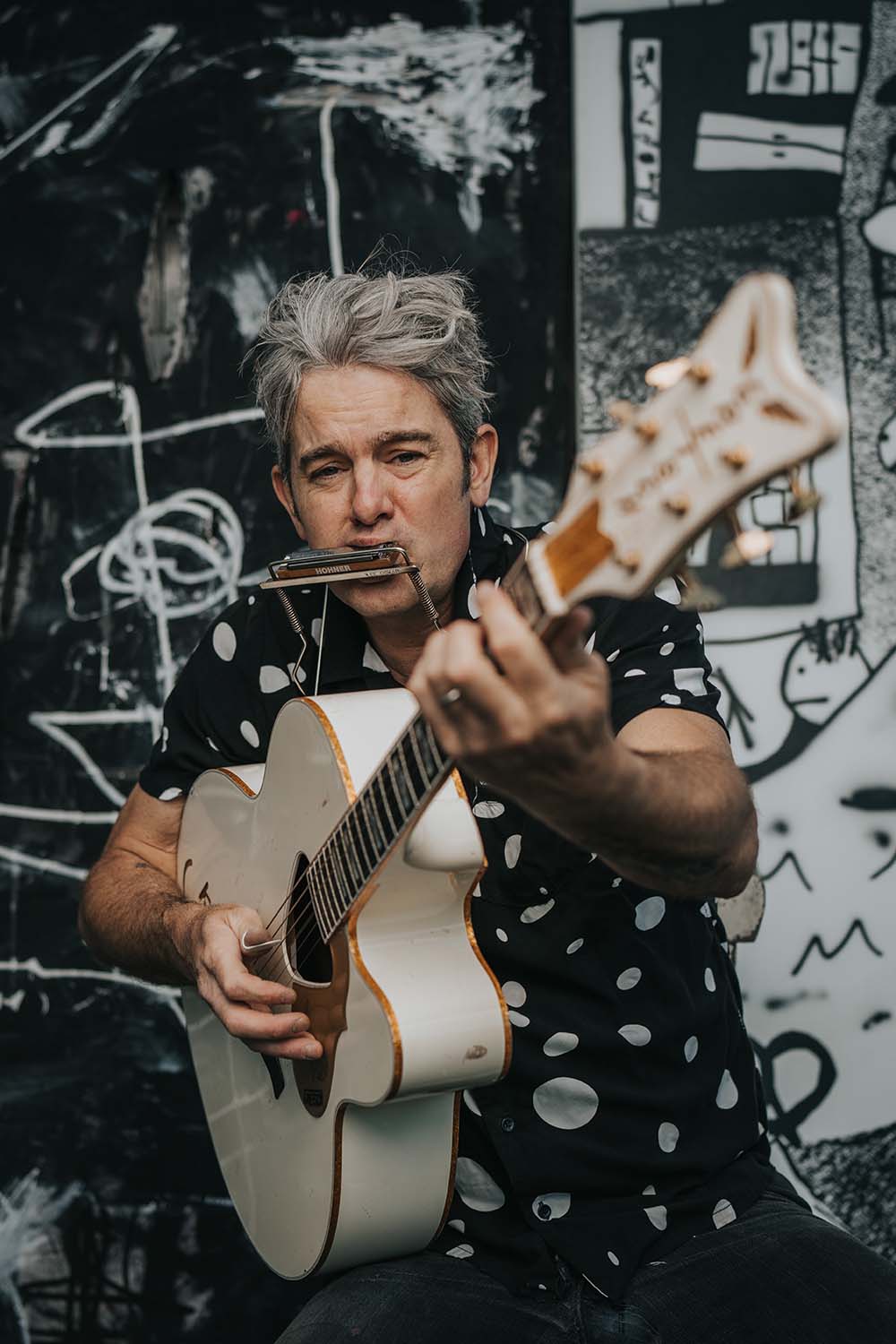 A man with gray hair wearing a polka dot shirt plays an acoustic guitar and a harmonica in front of a black and white abstract mural at the Cape Cod Arts Festival.