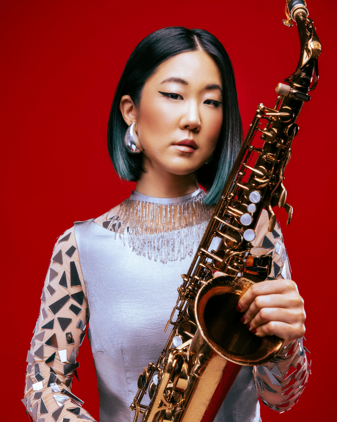 A woman with straight black hair holds a gold saxophone against a red background, wearing a silver dress with geometric patterns and large metallic earrings—ready to perform at the Cape Cod Jazz Festival.