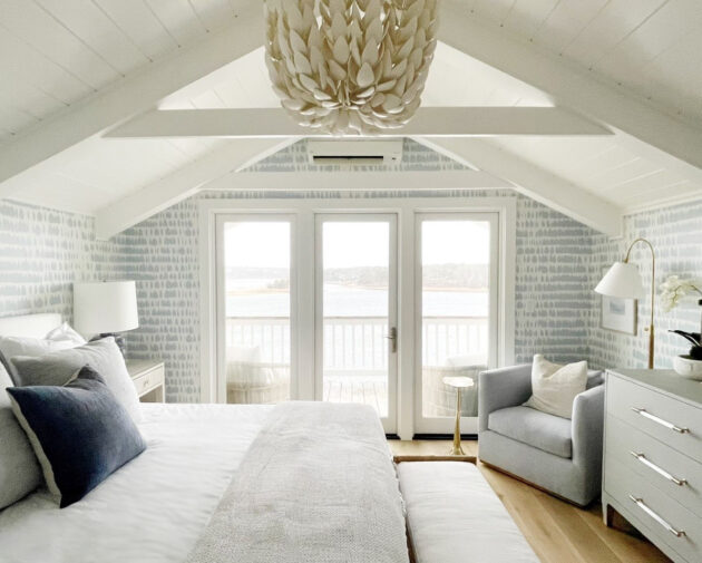 A cozy, light-filled bedroom features a bed, armchair, dresser, and a large chandelier under a vaulted ceiling, with glass doors opening to a balcony overlooking a water view.