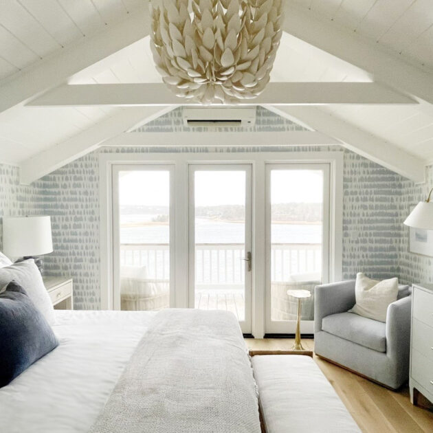 A cozy, light-filled bedroom features a bed, armchair, dresser, and a large chandelier under a vaulted ceiling, with glass doors opening to a balcony overlooking a water view.