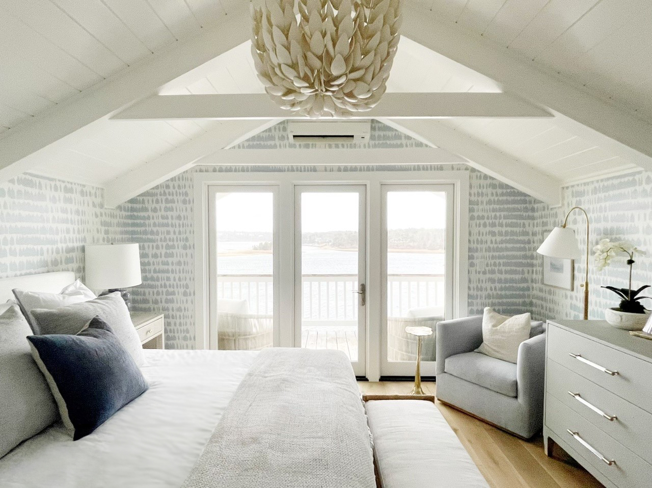 A cozy, light-filled bedroom features a bed, armchair, dresser, and a large chandelier under a vaulted ceiling, with glass doors opening to a balcony overlooking a water view.