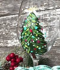 A Merry and Bright wine glass painted with a decorated Christmas tree—complete with ornaments and a star—stands before a rustic wooden background adorned with festive greenery and red berries; perfect for holiday painting lovers.