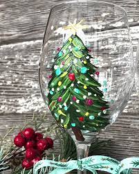 A Merry and Bright wine glass painted with a decorated Christmas tree—complete with ornaments and a star—stands before a rustic wooden background adorned with festive greenery and red berries; perfect for holiday painting lovers.
