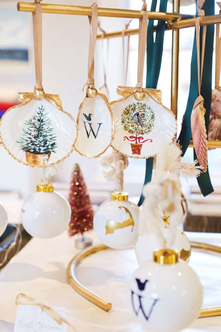 Decorative Christmas ornaments, including painted shells with trees and the letter "W," and white and gold baubles, hang from a gold display stand at the Merry Market annual event.