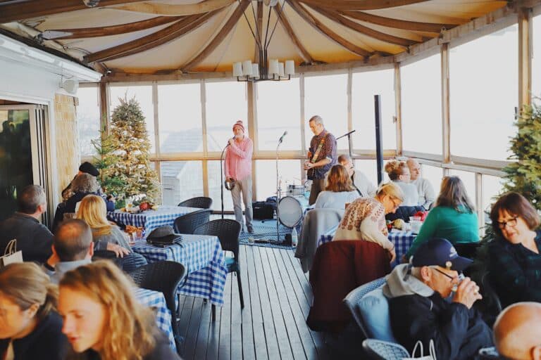 A small band performs on a covered patio during the Merry Market while diners sit at tables with blue checkered tablecloths, eating and conversing.