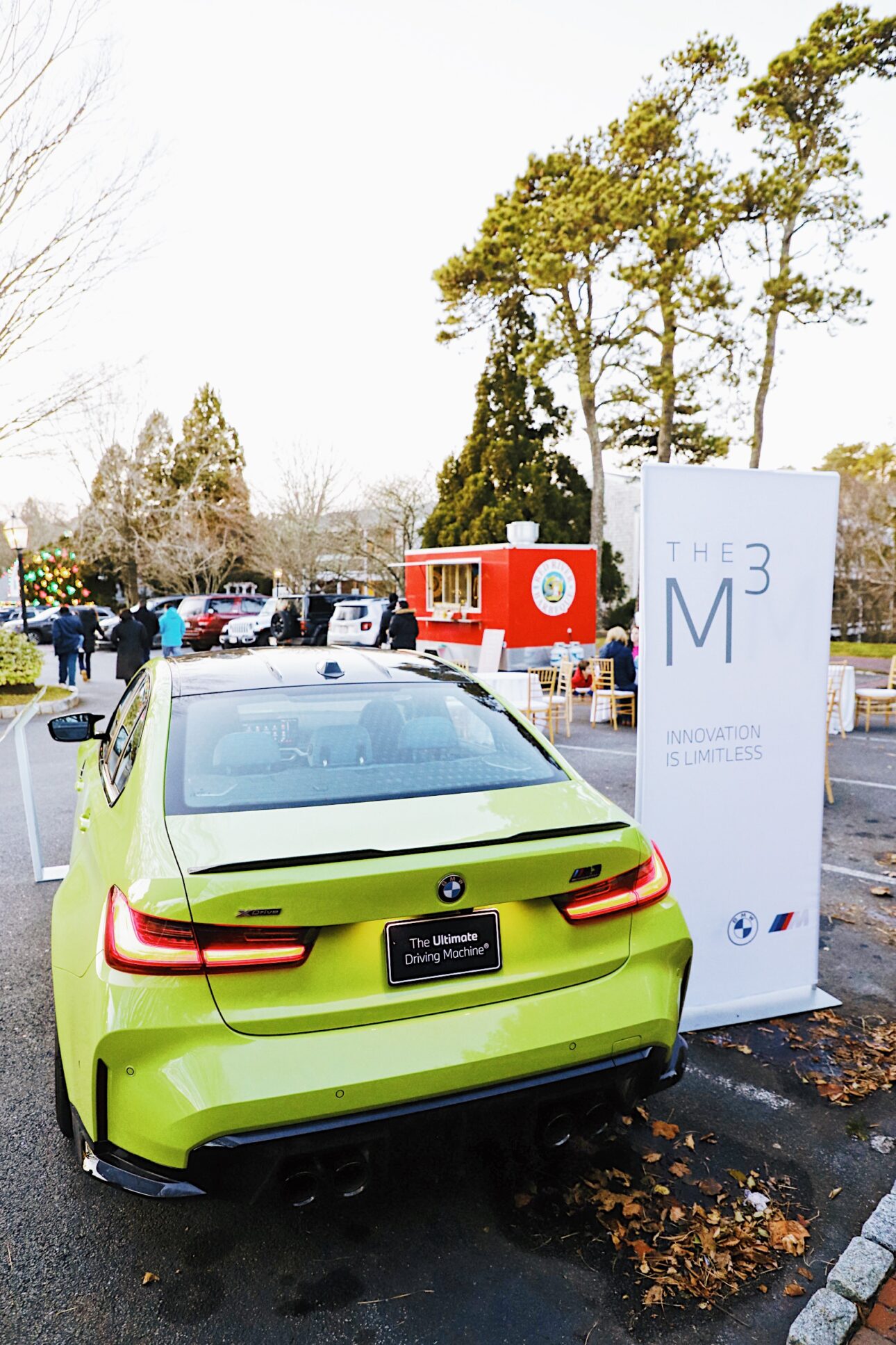 A green BMW M3 is parked near a promotional sign and food trucks at an outdoor event with people in the background, reminiscent of gatherings at Wequassett Resort and Golf Club or after a round at a Cape Cod golf course.