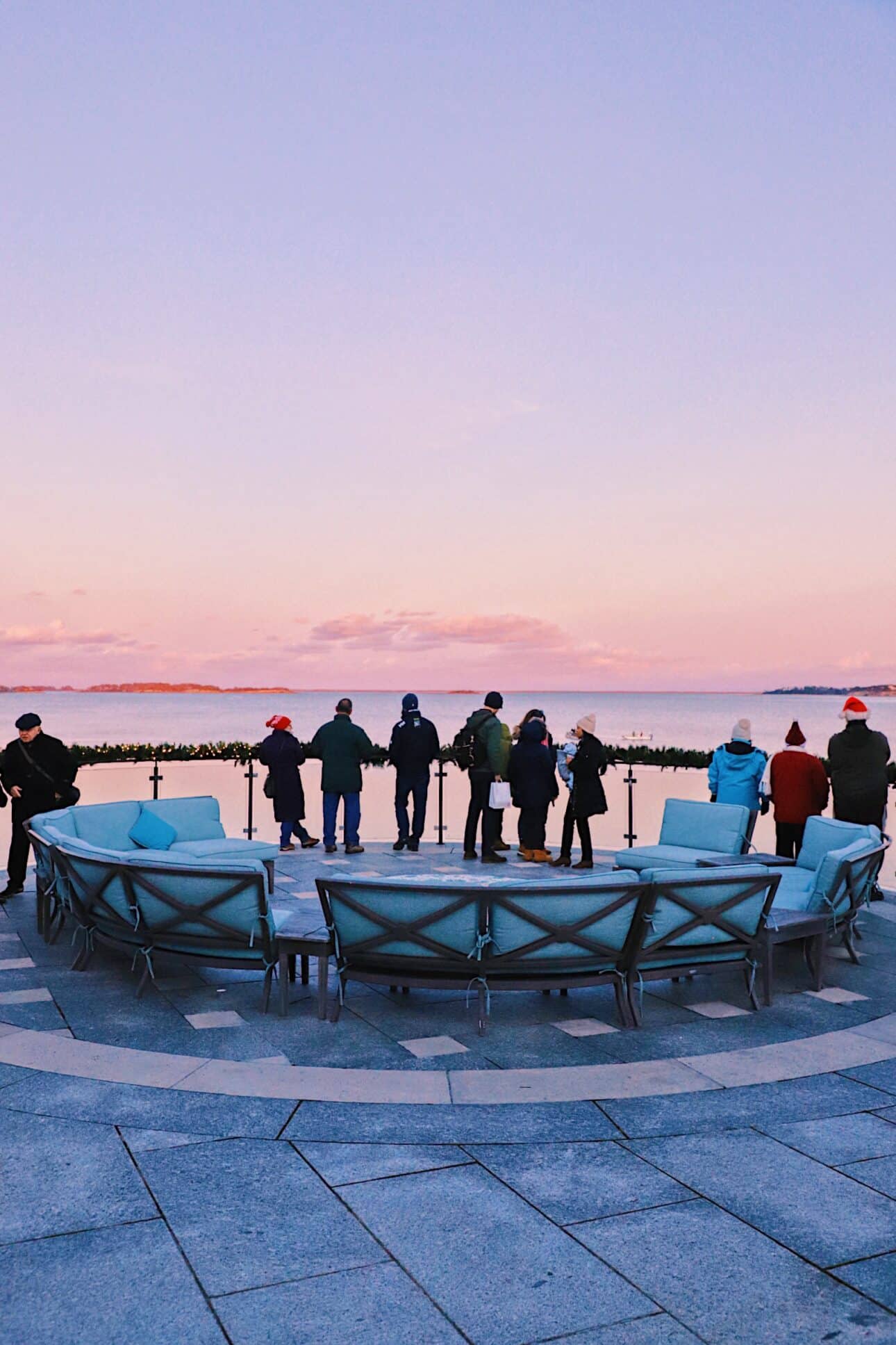 A group of people stand by a waterfront railing at sunset, with empty blue chairs arranged in a circle in the foreground, suggesting the anticipation of an annual event or fireworks display.