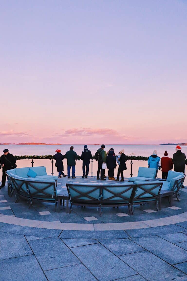A group of people stand by a waterfront railing at sunset, with empty blue chairs arranged in a circle in the foreground, suggesting the anticipation of an annual event or fireworks display.