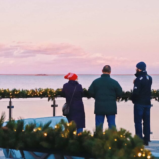 Three people stand by a decorated railing with string lights, looking out over a calm body of water at sunset, enjoying the peaceful scene after the Merry Market and tree lighting festivities.