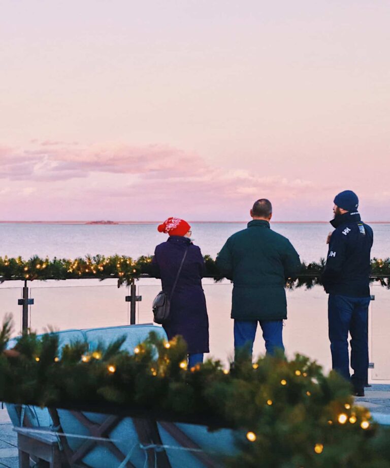 Three people stand by a decorated railing with string lights, looking out over a calm body of water at sunset, enjoying the peaceful scene after the Merry Market and tree lighting festivities.