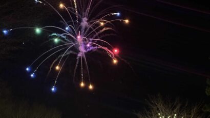 Colorful fireworks burst in the night sky above a building and trees decorated with white holiday lights during the Merry Market Tree Lighting, as people and cars gather below.