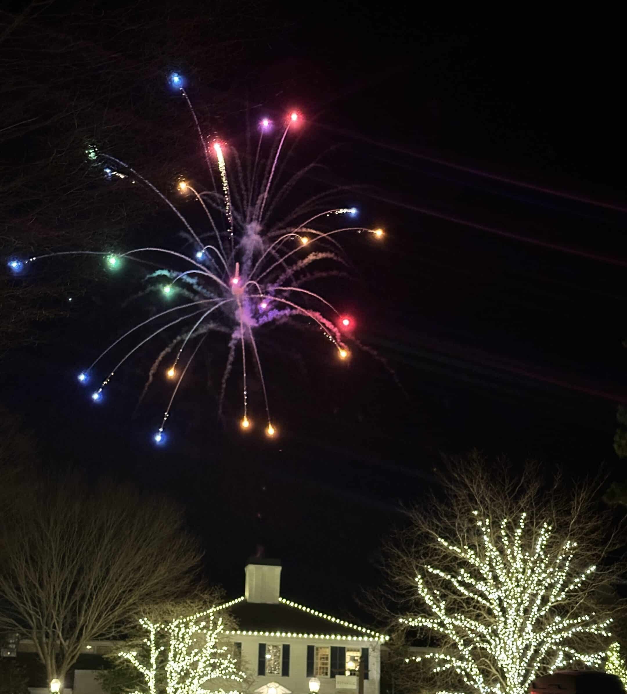 Colorful fireworks burst in the night sky above a building and trees decorated with white holiday lights during the Merry Market Tree Lighting, as people and cars gather below.