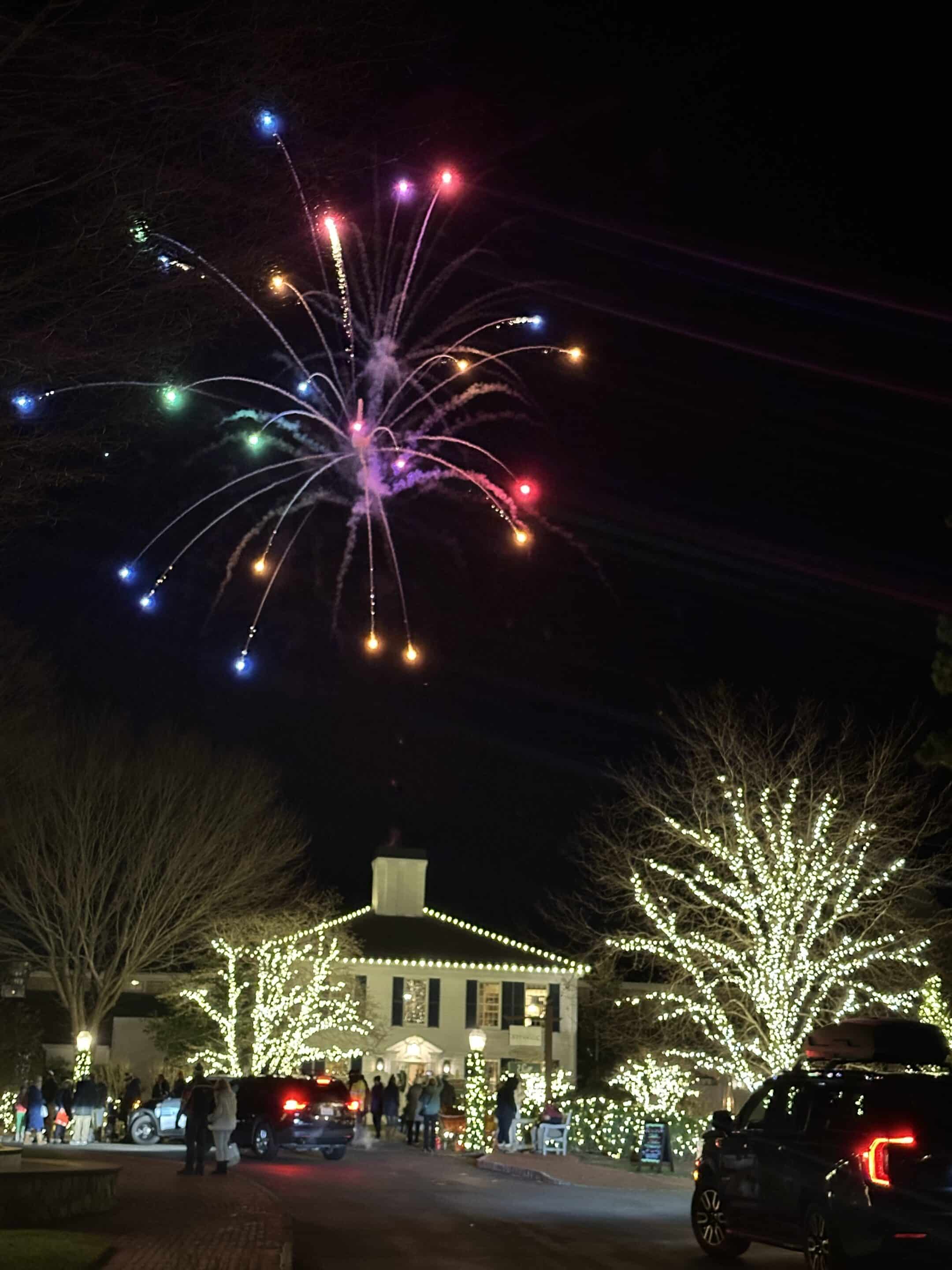 Colorful fireworks burst in the night sky above a building and trees decorated with white holiday lights during the Merry Market Tree Lighting, as people and cars gather below.